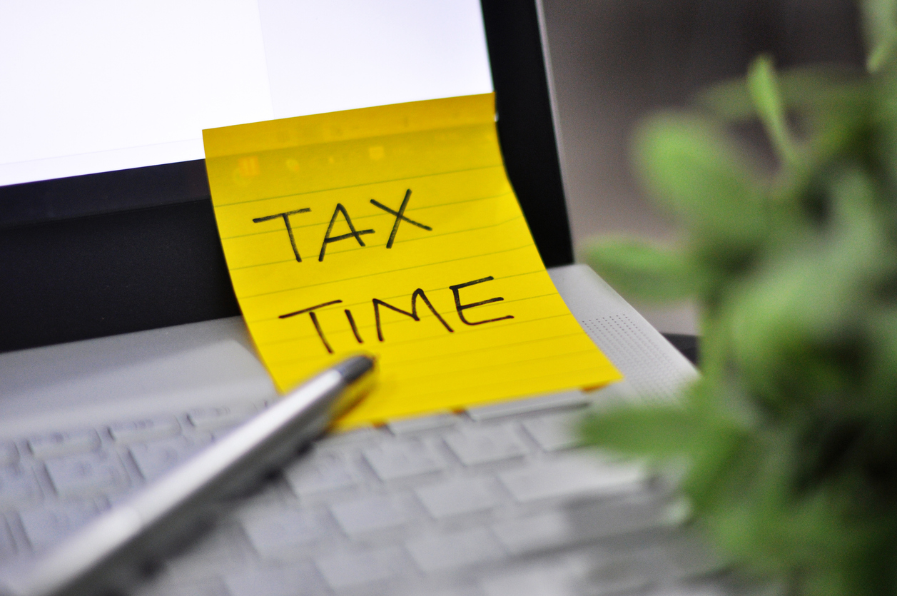 Tax Time: Horizontal top right view of an office laptop background with a metallic pen Tax Time note pad reminder green tree foregrounds - image